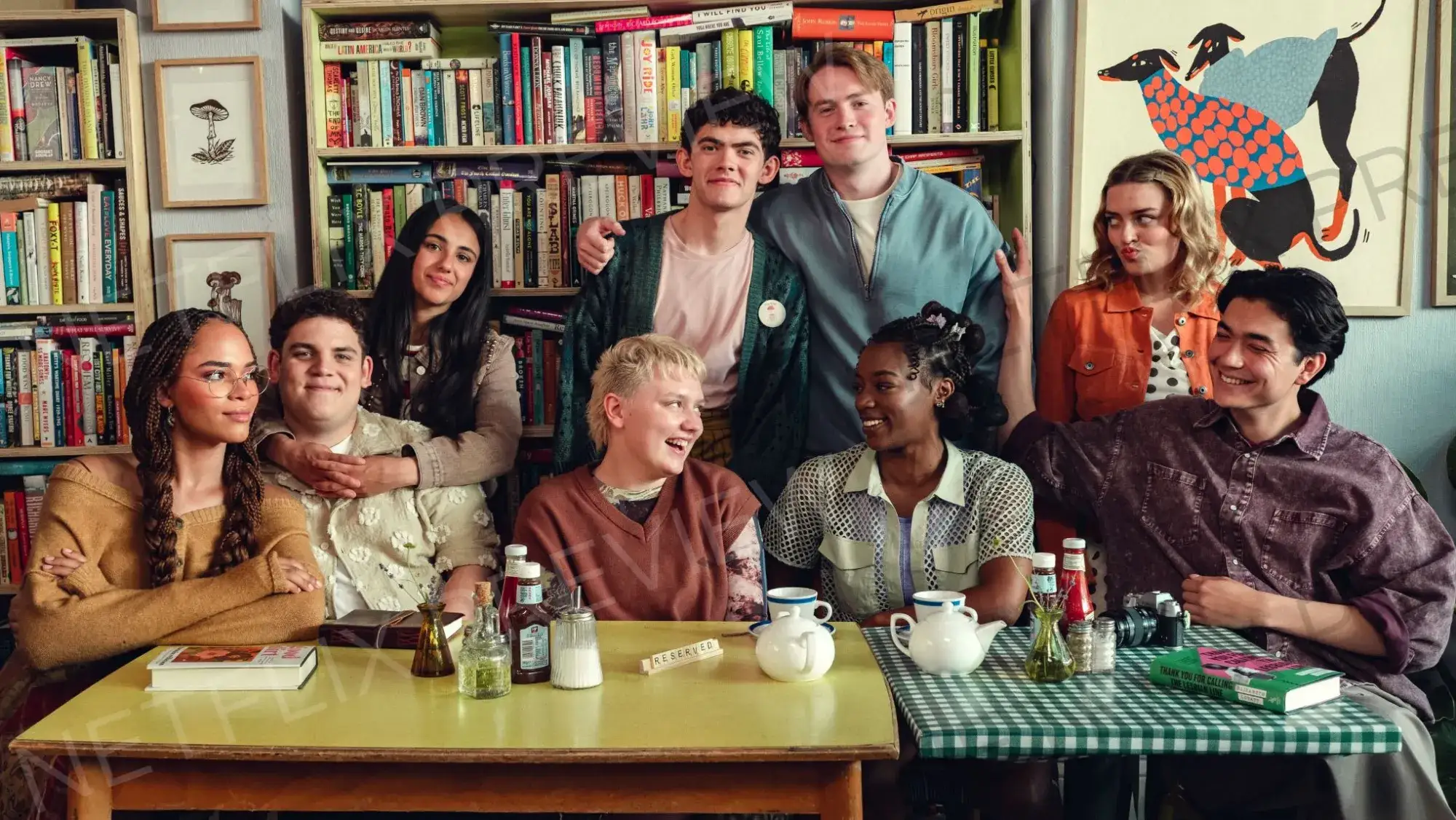 The cast of Heartstopper gathered around a table in a cozy bookshop cafe, smiling and laughing together